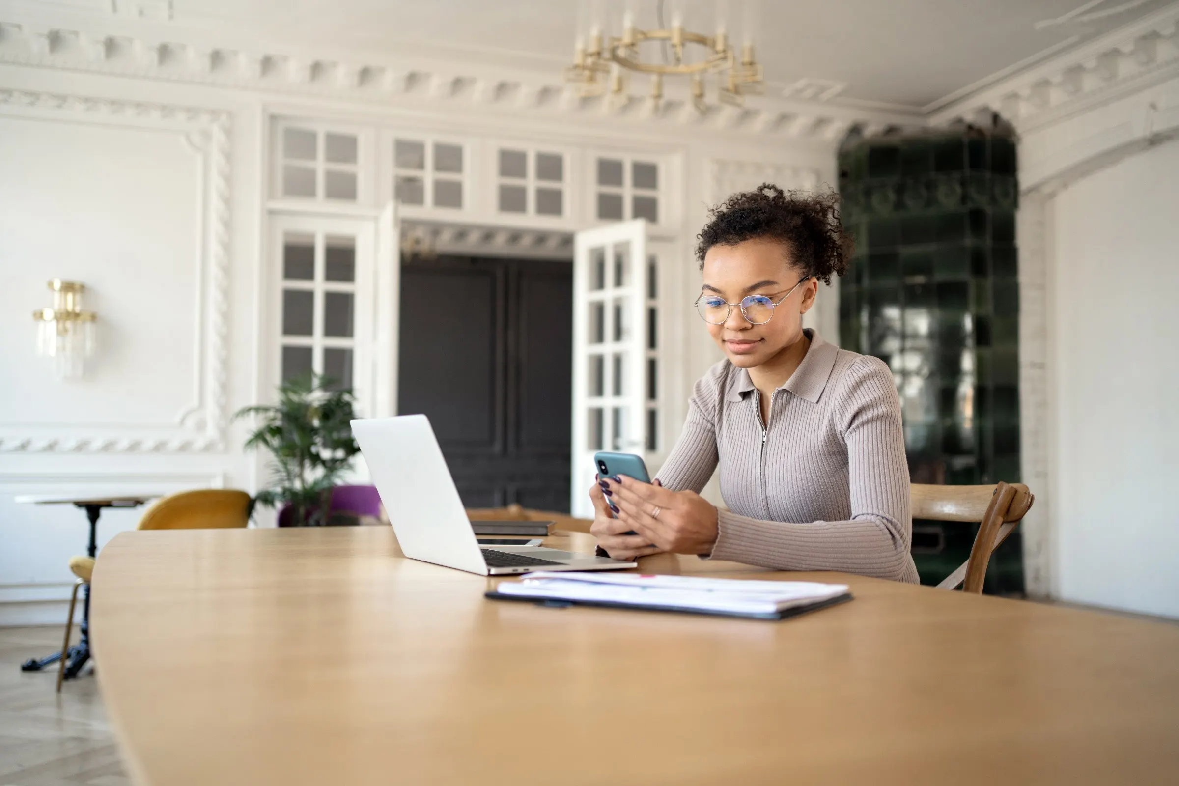 Femme qui consulte son téléphone devant un ordinateur portable, dans une pièce ancienne à moulures