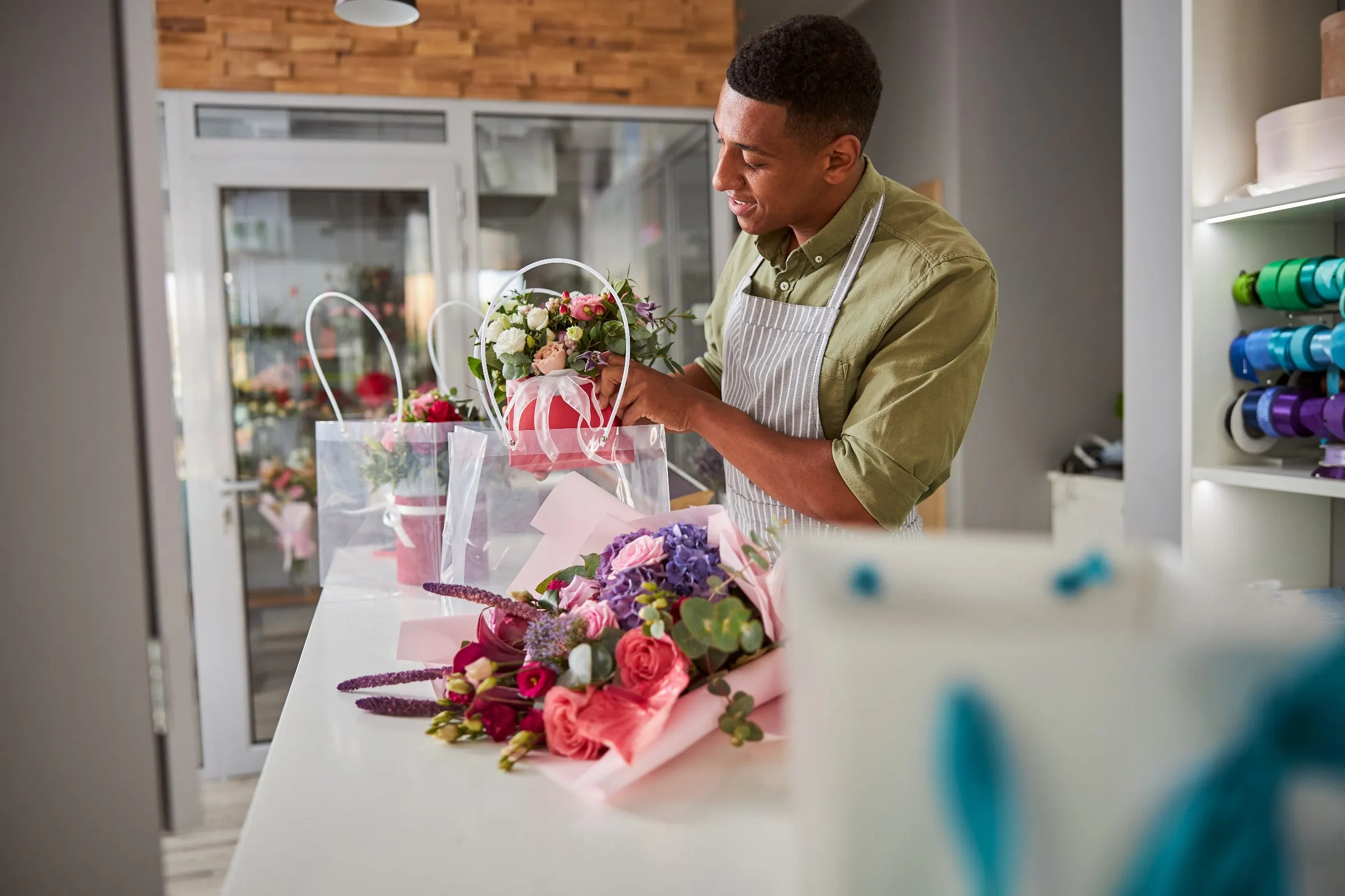 Fleuriste en tablier qui glisse un bouquet dans un sac transparent, sur le comptoir de sa boutique