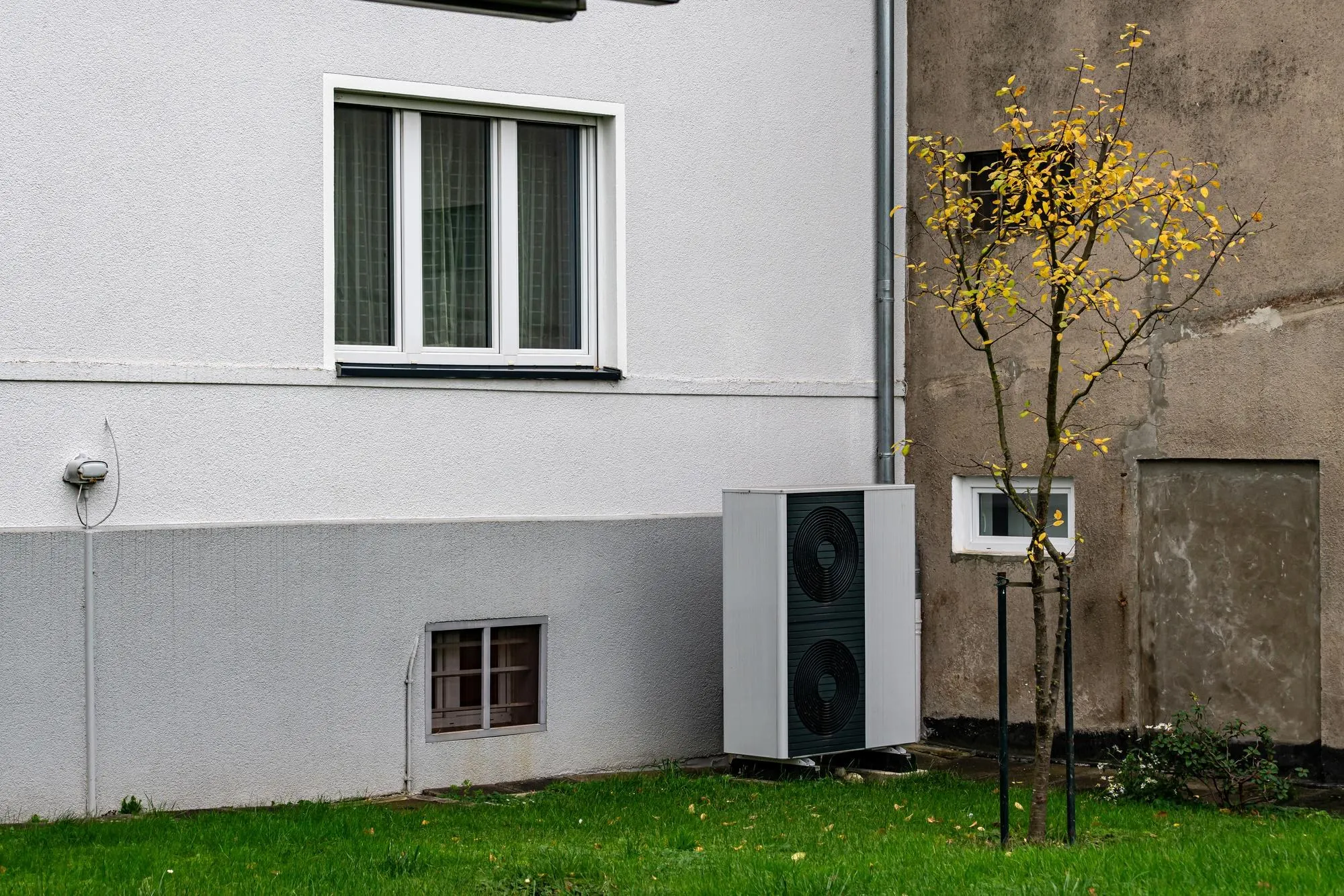 Unité extérieure d'une pompe à chaleur posée sur la pelouse, au pied du mur crépi d'une maison